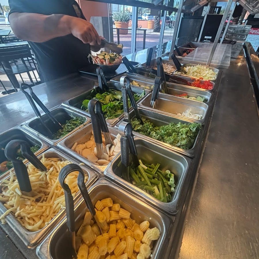 A staff member building a stir-fry bowl at the fresh ingredient line inside Blazin' Mongolian BBQ at The Pavilions at Talking Stick in Scottsdale