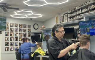 pavilions-barbershop-scottsdale-phil-master-barber Master barber Phil Malayev cutting a client's hair at The Pavilions Barbershop in Scottsdale, with a memorabilia wall and vintage chairs in the background